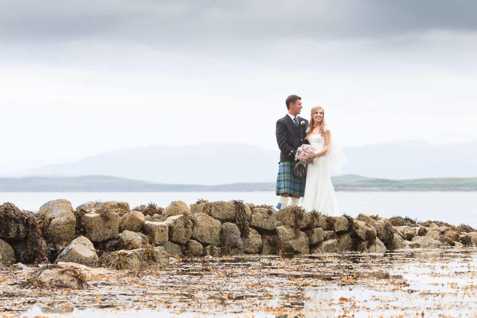 isle-of-skye-dunvegan-wedding a bride and groom stand on rocks by the waters edge in a scottish wedding photograph from the isle of skye